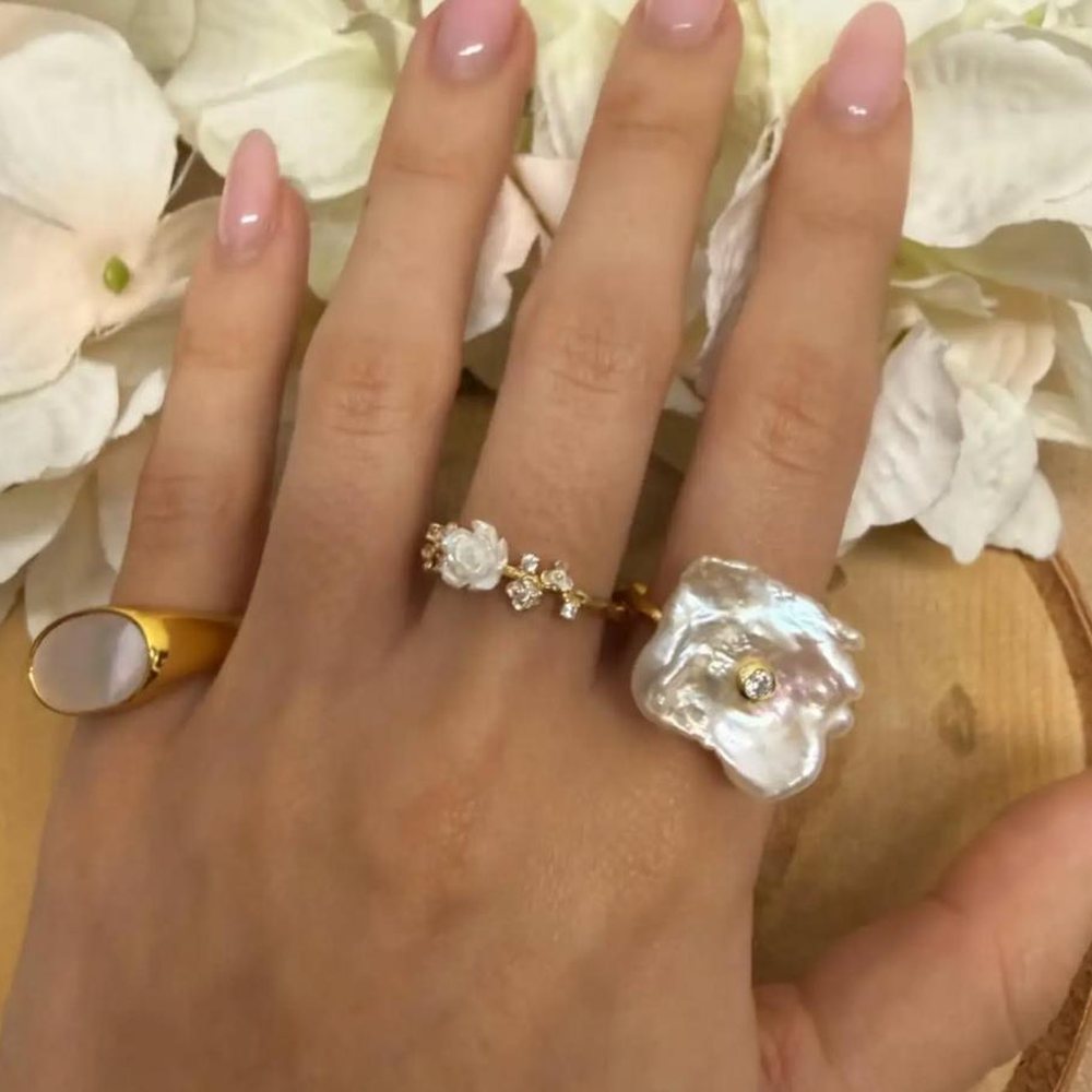 Mão com três anéis: um selo em ouro com madrepérola, um anel floral com zircónias e um anel de pérola barroca
