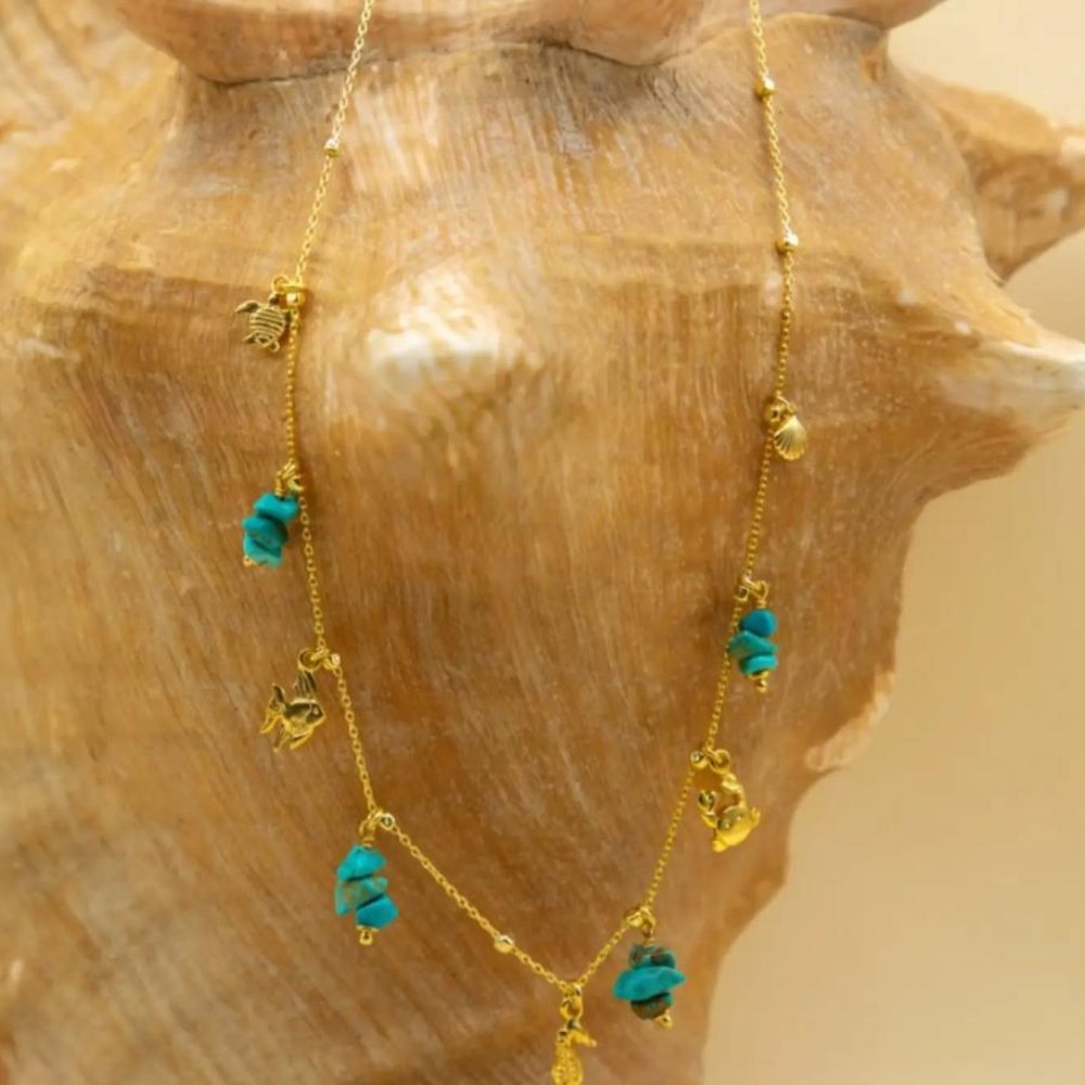 Colar dourado com pedras de turquesa e pendentes com motivos marinhos, sobre um búzio